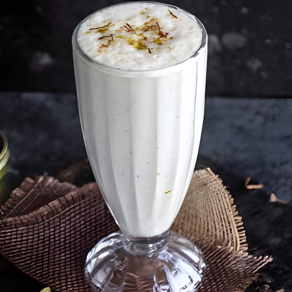 A tall glass of creamy white lassi, topped with saffron threads, on a rustic brown mat.