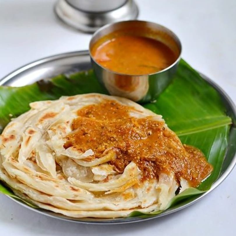 Flaky Indian parotta bread topped with a rich brown curry, served on a banana leaf.
