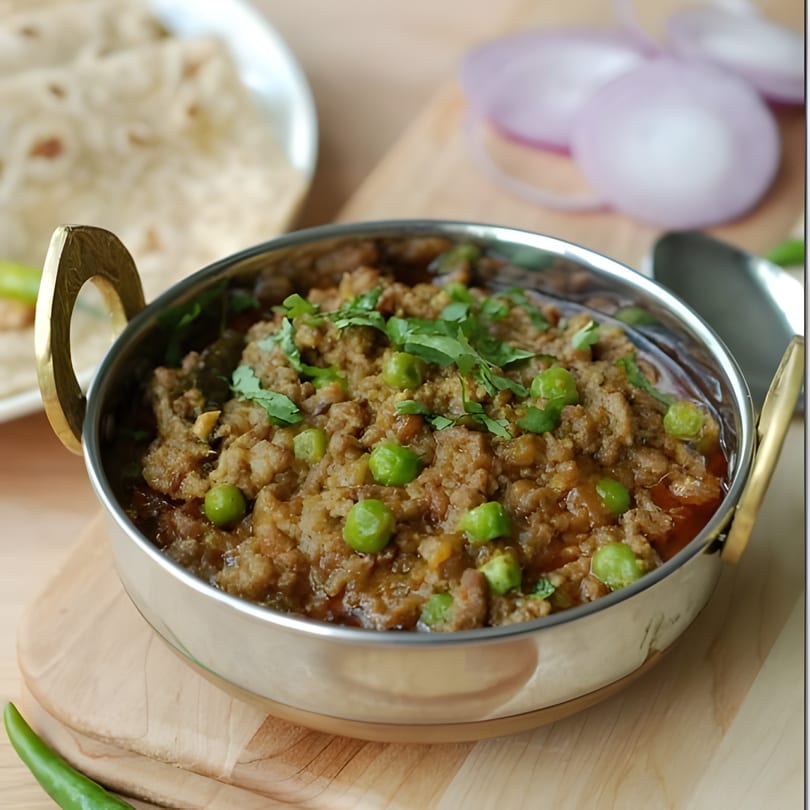 A stainless steel bowl of Indian keema matar, garnished with cilantro and peas, on a wooden board.
