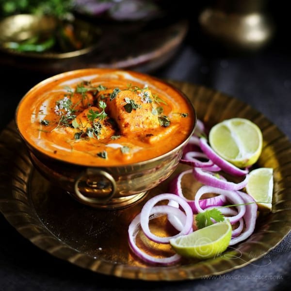 A brass bowl of paneer butter masala, garnished with herbs, served with red onion and lime.